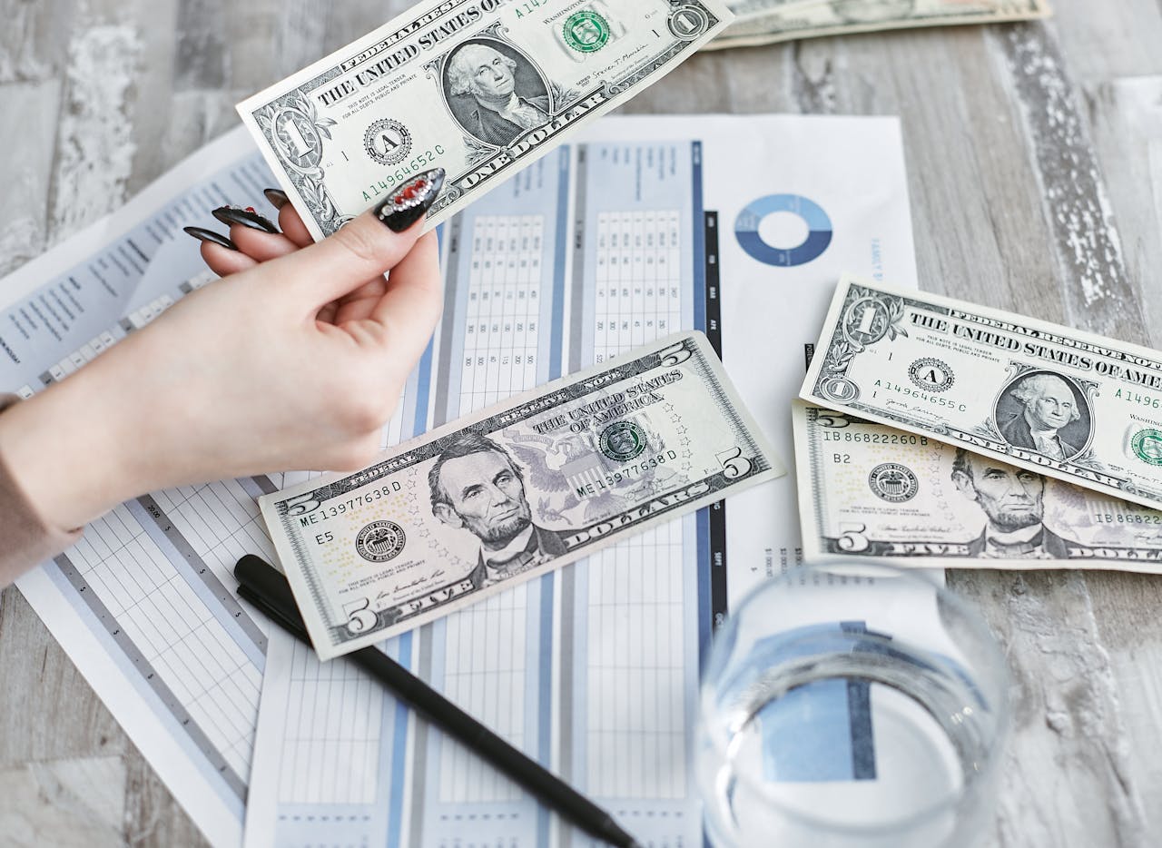 Close-up of a hand holding a dollar bill over financial documents and charts on a wooden table.